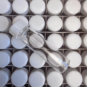 May include: A clear glass bottle with a white screw-on cap positioned diagonally across a grid of identical white bottle caps. The bottle is transparent and cylindrical, featuring a ribbed white plastic lid that matches the caps in the background.