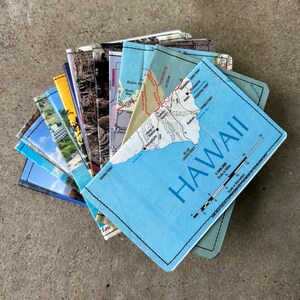 May include: A stack of colourful travel guides with maps of Hawaii. The top guide is blue with the word "HAWAII" in large white letters and a map of the islands. The guides are arranged in a fan shape.