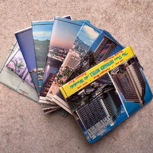 May include: A collection of handmade postcard wallets, each featuring a unique vintage travel scene. The wallets are made from sturdy fabric and showcase images of cityscapes, sunsets, and tropical locations. One wallet displays the text "The Four OUTRIGGER HOTELS At Waikiki".