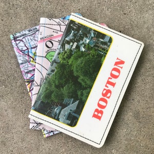 May include: A white notebook with a black stitched border and the word "BOSTON" in red letters. The notebook has a photo of an aerial view of a city with trees and buildings. The notebook is on top of a folded map of a city with the words "MEDFORD", "MALDEN", "BEACON AVE", "SOMERVILLE", "CAMBRIDGE", "GRIGGS", "ROAD", "RD", and "M" visible.