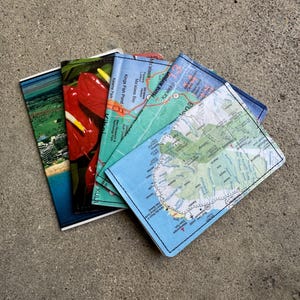 May include: A stack of fabric coasters featuring various designs. One coaster displays a scenic aerial view, another showcases red flowers, and others feature detailed map prints with place names like Hana Airport and Ma'alaea Bay. The coasters are square and have black stitching.
