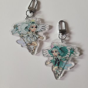 May include: Two clear acrylic keychains featuring a winged character in a chibi art style. One character wears a white outfit with gold accents, while the other wears a teal outfit with dark details. Both figures hold a staff and have silver clasps.