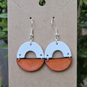 May include: A pair of white and orange leather earrings with a half-circle design. The earrings are hanging from silver hooks.