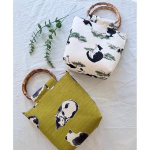 May include: Two tote bags with bamboo handles. One bag is olive green with a black and white cat pattern. The other bag is off-white with a black and white cat and pine tree pattern. Both bags are square-shaped.