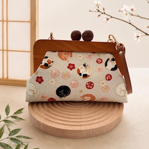 May include: A wooden-framed clutch bag with a light fabric patterned with sleeping cats and floral designs in red, pink, and white. It has a brown leather strap and two dark brown spherical closures.