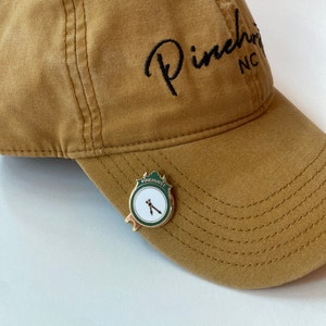 May include: A brown baseball cap with the text "Pinehurst NC" embroidered on the front. A gold-colored pin with a green and white design is attached to the cap. The pin features the text "Pinehurst" and a clock face with two hands.