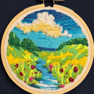 Pode incluir: Arte de bordado em aro que representa uma paisagem com um céu azul, nuvens brancas, colinas verdes, um campo amarelo e um rio azul serpenteando pelo centro.