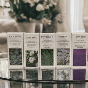 Könnte beinhalten: Fünf kleine Flaschen mit ätherischem Öl mit Etiketten für Eukalyptus, Gardenie, Pfefferminze, Freesie und Lavendel. Jede Flasche hat ein weißes Etikett mit schwarzem Text und einem kleinen Bild der entsprechenden Blume oder Pflanze.