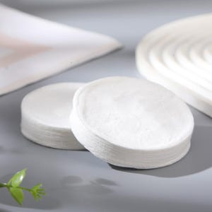 Può includere: Due pile di dischetti di cotone rotondi e bianchi sono a fuoco, con alcuni altri oggetti bianchi sullo sfondo. I dischetti sono morbidi e soffici, suggerendo un prodotto per la cura della pelle o cosmetico. L'immagine è ben illuminata.