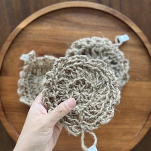 Puede incluir: Tres posavasos redondos beige, tejidos a ganchillo, con un borde en bucle y un pequeño lazo para colgar. Los posavasos están hechos de fibra natural y se exhiben en una bandeja de madera.