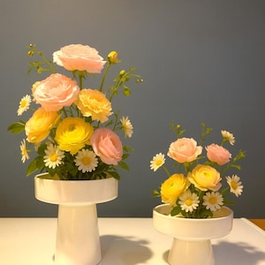 Puede incluir: Dos jarrones de pedestal de cerámica blanca con arreglos florales. Los arreglos presentan rosas amarillas y rosas, pequeñas margaritas blancas y tallos verdes. Los jarrones están sobre una superficie blanca sobre un fondo azul grisáceo.