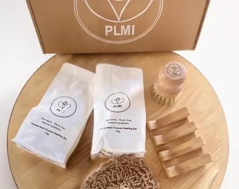 May include: A zero-waste cleaning kit with a cardboard box, two white packages of soap, a wooden dish brush, a woven scrub, and a wooden soap dish. The packages read "Oatmeal Multi Purpose Washing Bar 150g". The box has the logo "PLMI".