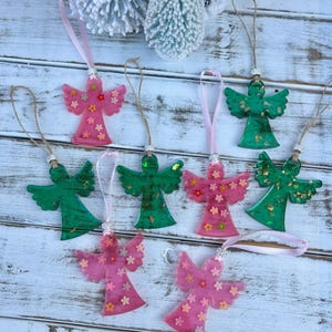 May include: A collection of handmade angel ornaments in shades of pink and green, adorned with small flower accents. Each ornament has a ribbon or twine for hanging. The angels are displayed on a rustic, white-washed wooden surface.