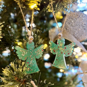 May include: Two green angel ornaments with glitter and red accents hang from twine strings. The angel wings are spread, and the ornaments are suspended in front of a Christmas tree.