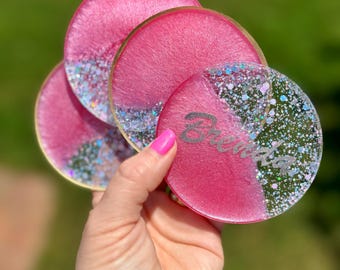 May include: Set of four pink and gold glitter resin coasters with a gold rim. The coasters have a white glitter design and the word "Believe" is written on one of the coasters.