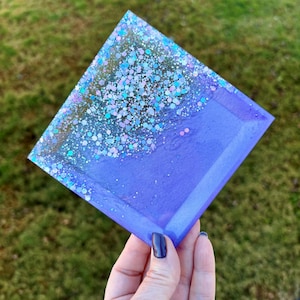 May include: A square purple resin coaster with iridescent glitter embedded in the resin. The glitter is concentrated at the top of the coaster and fades to a solid purple at the bottom.