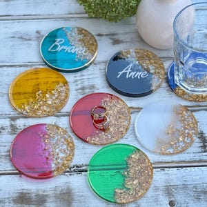 May include: A collection of colorful, round resin coasters with gold leaf accents. Some coasters have names written on them, and one holds a ring. Colors include teal, yellow, red, pink, green, white, and navy.