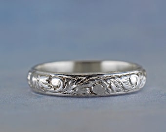 May include: A silver band ring with an intricate floral design. The band is smooth and polished, and the floral design is raised and detailed.