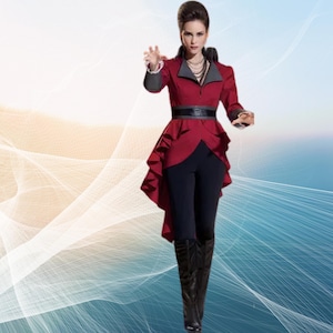 May include: A woman wearing a red and black jacket with a ruffled hem and a black belt. She is standing in front of a blue and white background.