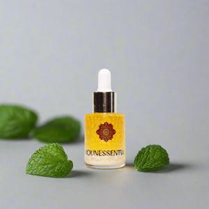 May include: A small, yellow glass bottle with a white dropper and gold accents, labeled "NOUNESSENTIAL". The bottle is surrounded by green leaves on a gray background. The bottle has a decorative floral design.