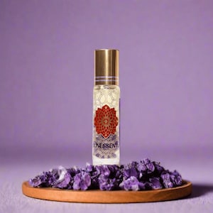 Könnte beinhalten: Eine Roll-on-Flasche mit ätherischem Öl, mit goldener Kappe und einem rot-goldenen Design, beschriftet mit "UNESSENT". Die Flasche steht auf einem Holztablett mit lila Kristallen, vor einem lila Hintergrund.