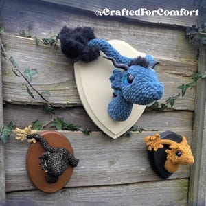Puede incluir: Tres cabezas de dragón de ganchillo montadas en placas de madera. Los dragones son azules, negros y naranjas. Las placas son blancas, marrones y negras. La imagen incluye el texto "Crochet Pattern".