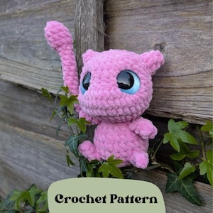 May include: Pink crocheted amigurumi toy with large blue eyes, resembling a cartoon character. The toy is perched on a wooden fence post with green vines growing around it. The text "Crochet Pattern" is written on a green label below the toy.
