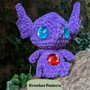 Puede incluir: Juguete amigurumi de crochet morado con detalles de gemas azules y rojas. El juguete está sentado en una rama de árbol. Patrón de crochet.