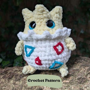 May include: A crocheted plush toy of a Pokemon character,  Egg-shaped with a white body, red and blue accents, and a yellow head with blue eyes. The toy is sitting on a tree branch. The text "Crochet Pattern" is visible below the toy.