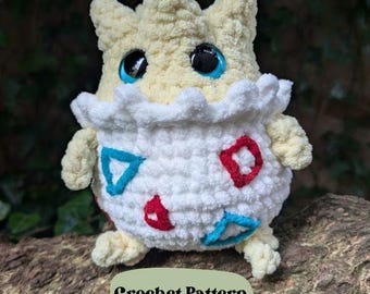 Puede incluir: Un juguete de peluche tejido a crochet de un personaje de Pokemon, con forma de huevo, cuerpo blanco, detalles rojos y azules, y cabeza amarilla con ojos azules. El juguete está sentado en una rama de árbol. El texto "Crochet Pattern" es visible debajo del juguete.