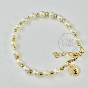 May include: A delicate gold bracelet with a single initial charm and a strand of white pearls.
