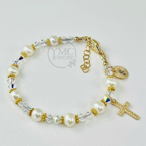 May include: Gold bracelet featuring white pearls, clear crystals, and a gold cross charm. The bracelet has a gold clasp and a small gold disc with the letter "S" on it.