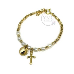 May include: Gold bracelet with white pearls, a gold cross charm, and a gold initial charm with the letter "S".