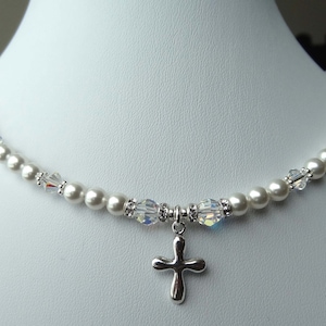 May include: A delicate white pearl necklace with a small silver cross pendant. The necklace is accented with clear crystal beads.
