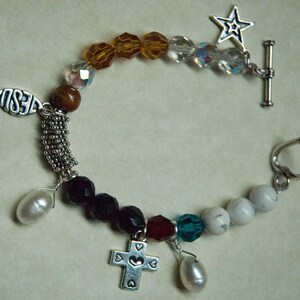 May include: A silver charm bracelet with a variety of charms, including a fish with the word "Jesus" on it, a cross, a heart, a star, and a pearl. The bracelet is made of silver beads and crystals.