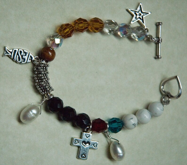 May include: A silver charm bracelet with a variety of charms, including a fish with the word "Jesus" on it, a cross, a heart, a star, and a pearl. The bracelet is made of silver beads and crystals.