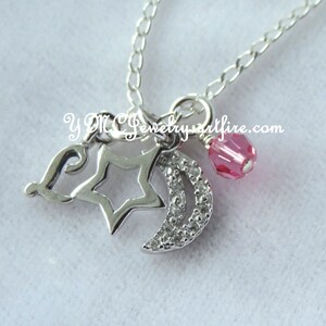 May include: A silver necklace with a charm featuring a crescent moon, a star, and a letter L. The charm is accented with small clear crystals. A pink crystal bead hangs from the charm.