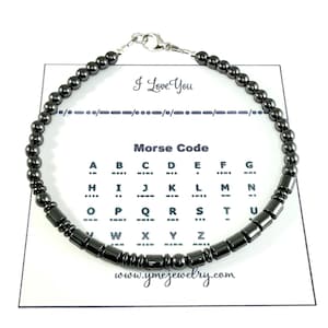 May include: A black beaded bracelet with a silver clasp, presented on a white card. The card displays "I Love You" in Morse code, alongside a Morse code alphabet chart. The website address www.ymcjewelry.com is also visible.