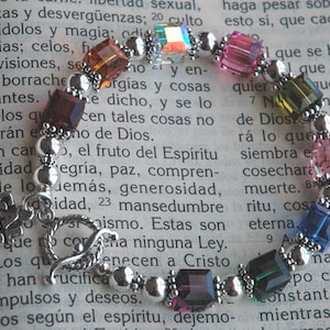 May include: A silver bracelet with a clasp closure features a variety of colorful square-shaped crystals. The crystals are in shades of pink, green, blue, purple, and orange. The bracelet is on a book with text in Spanish.