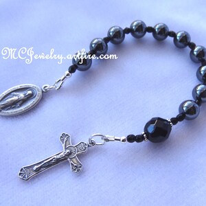 May include: A black rosary bracelet with a silver crucifix and a silver charm of the Virgin Mary. The beads are made of hematite and the center bead is black onyx.