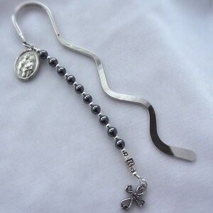 May include: A silver bookmark with a black beaded chain, a silver charm with a cross, and a silver charm with a religious image.
