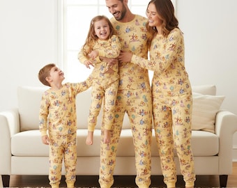May include: A family of four wearing matching yellow pajamas with a cartoon character print. The pajamas include long-sleeved tops and pants. The family is standing in a living room with a white couch and a window.