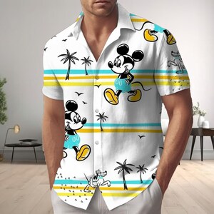 Könnte beinhalten: Weißes Button-Down-Hemd mit einem sich wiederholenden Muster aus Mickey Mouse, Palmen und Vögeln. Das Design umfasst gelbe und blaue horizontale Streifen. Das Hemd hat kurze Ärmel und einen klassischen Kragen.