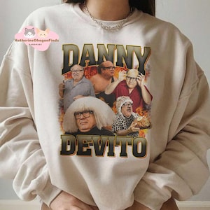 May include: A beige sweatshirt with a graphic print of Danny DeVito. The print features a collage of images of Danny DeVito in different roles, including Frank Reynolds from the show 'It's Always Sunny in Philadelphia'. The text 'Danny DeVito' is printed in large, bold letters below the images.
