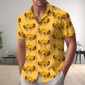 May include: Yellow button-down shirt featuring a repeating pattern of Simba's face from The Lion King. The short-sleeved shirt has a classic collar and white buttons. The shirt is made of a lightweight fabric.