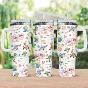 May include: Three white insulated tumblers with handles, lids, and straws. Each tumbler features a repeating pattern of Toy Story characters, stars, and the phrase "You've Got a Friend in Me". The tumblers are on a wooden surface.