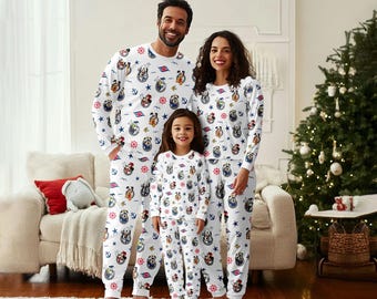 May include: A family of three wearing matching white pajamas with a nautical and cartoon dog print. The pajamas feature long sleeves and pants. The background includes a Christmas tree, a couch, and a rug.