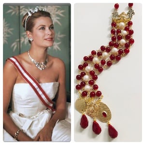 Puede incluir: Un brazalete de cuentas doradas y rojas con un dije colgante. El brazalete presenta cuentas rojas, perlas blancas y eslabones de cadena dorados. El dije es un colgante dorado con un diseño floral y tres cristales rojos en forma de lágrima.