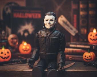 May include: A posable action figure of a character with a white mask, dark hair, and a black jumpsuit. The figure is seated, holding a small silver-colored knife in one hand. The mask features dark eye sockets and a neutral expression.