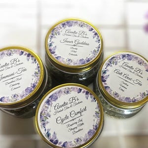 Puede incluir: Cuatro frascos de vidrio de t&eacute; de hierbas con tapas doradas. Cada frasco tiene una etiqueta con el texto "Auntie B's Herbal Teas & Tinctures" y un nombre de t&eacute; diferente. Las etiquetas tienen un dise&ntilde;o floral.